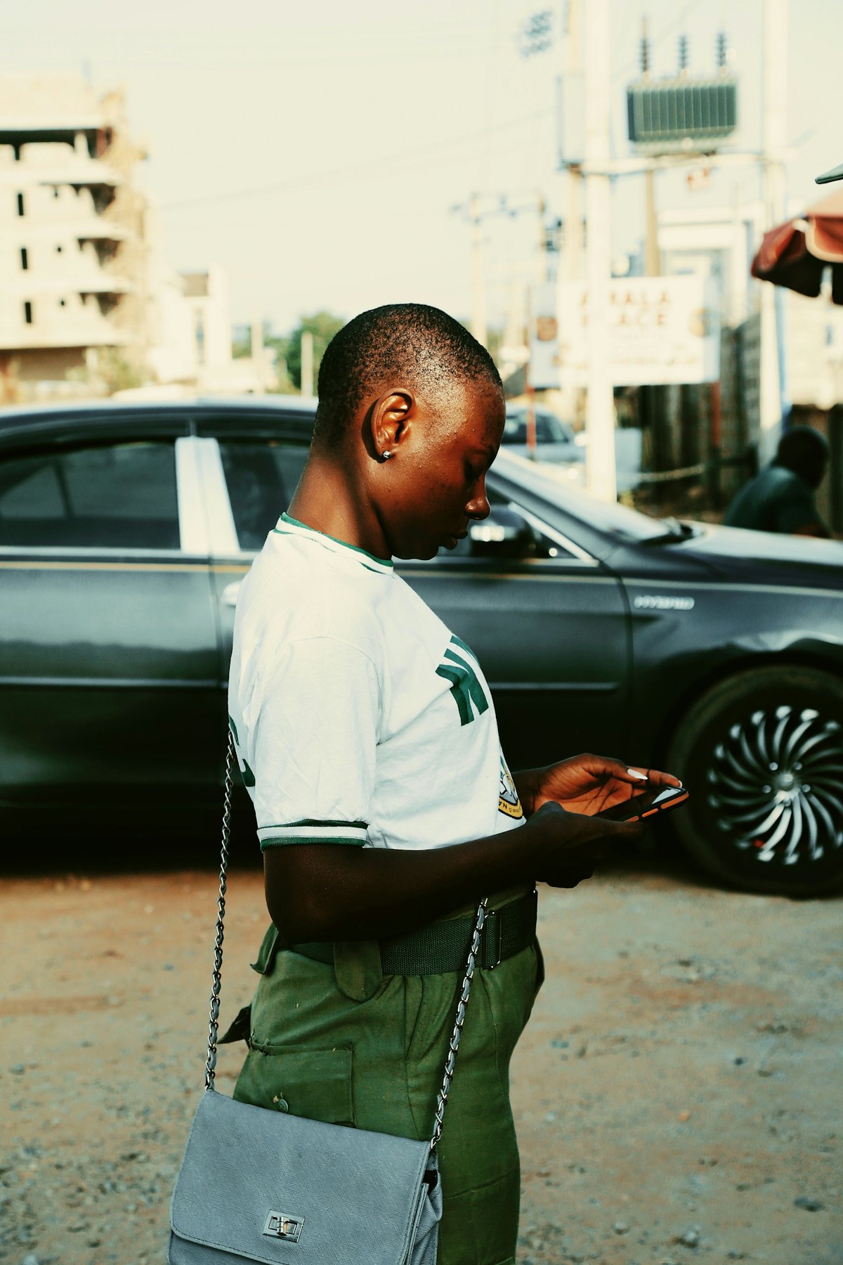A young person in a green and white uniform checking their phone on a street in Nigeria